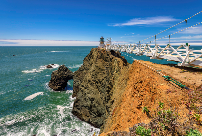 A lighthouse out on a rocky peninsula reached by a suspension bridge.